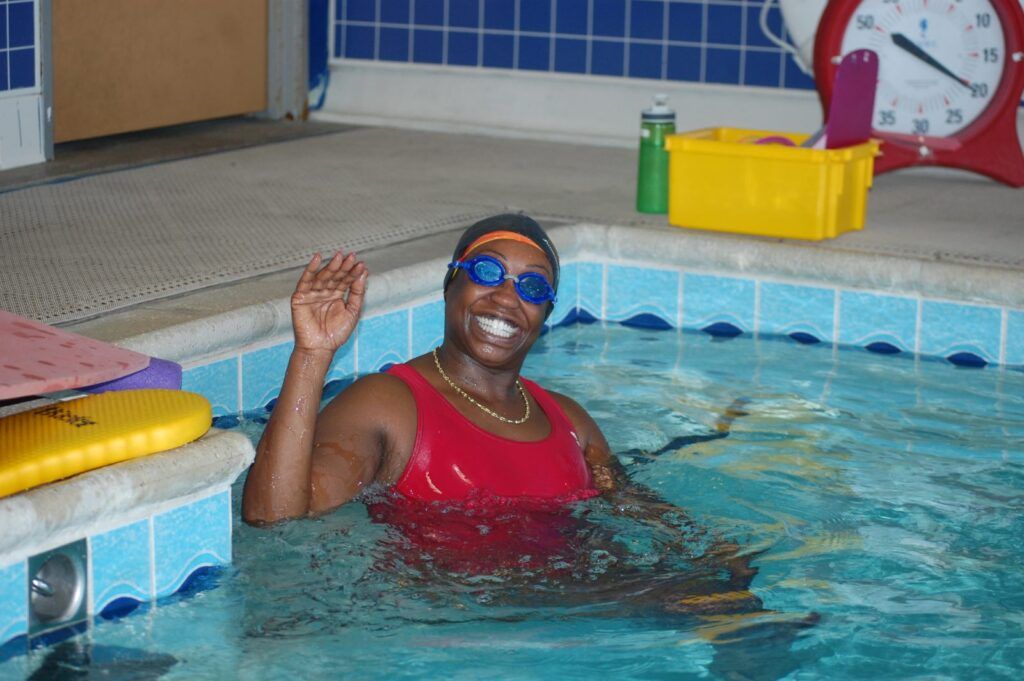 Adult swimmer smiling and waving while standing in a pool, wearing goggles and a red swimsuit, with swim equipment and a pool deck visible in the background.