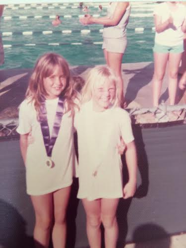 Two young girls standing side by side in front of a swimming pool, smiling and wearing oversized T-shirts, with one girl wearing a medal around her neck at what appears to be a childhood swim event.